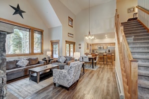 A spacious living area with a large sofa, patterned armchair, and a wooden coffee table. A staircase with wooden railings leads to an upper level.