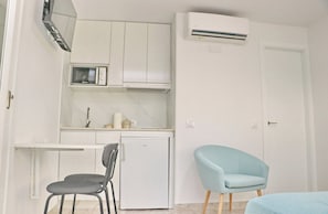 A compact kitchen with white cabinets, a microwave, a small refrigerator, and a light blue chair. - The little Loft    . (Conil de la Frontera)