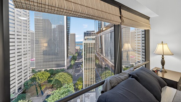 A modern hotel room with a large window offering a city view, a comfortable bed, a bedside table with a lamp, and a view of the surrounding urban landscape. - Chic Dt Honolulu Executive Condo 1 Bedroom Condo (Honolulu)