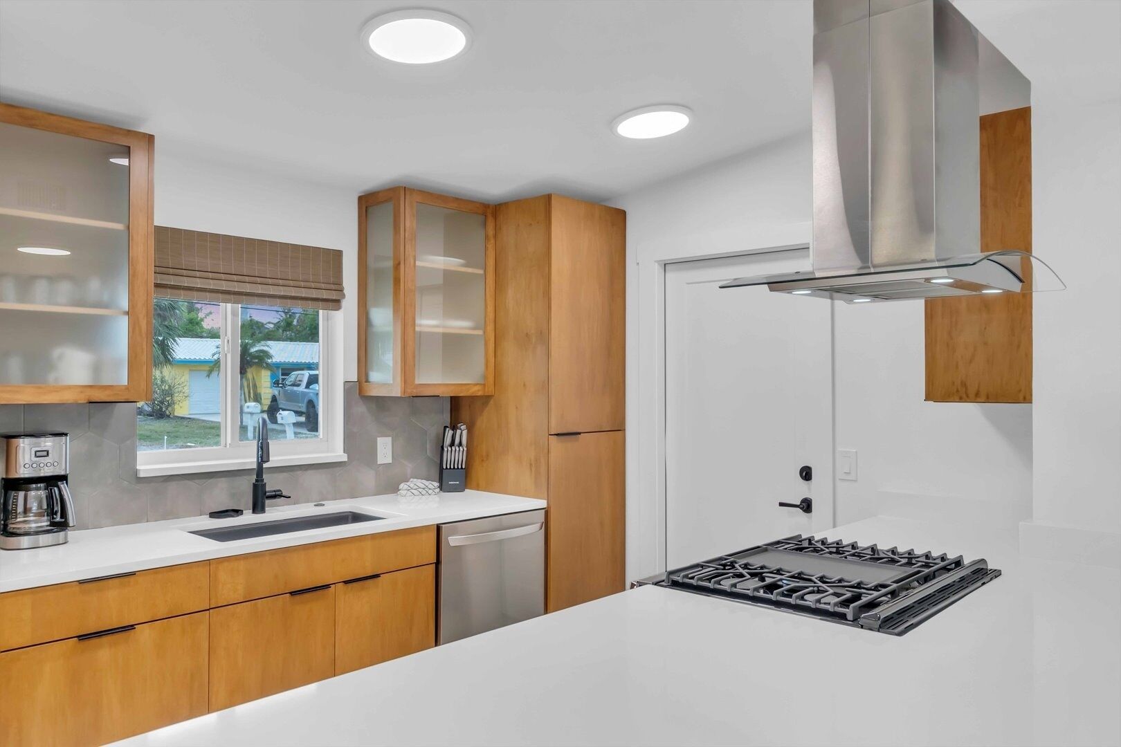 A modern kitchen with wooden cabinets, a stainless steel range hood, a dishwasher, and a window with a view of the outside.