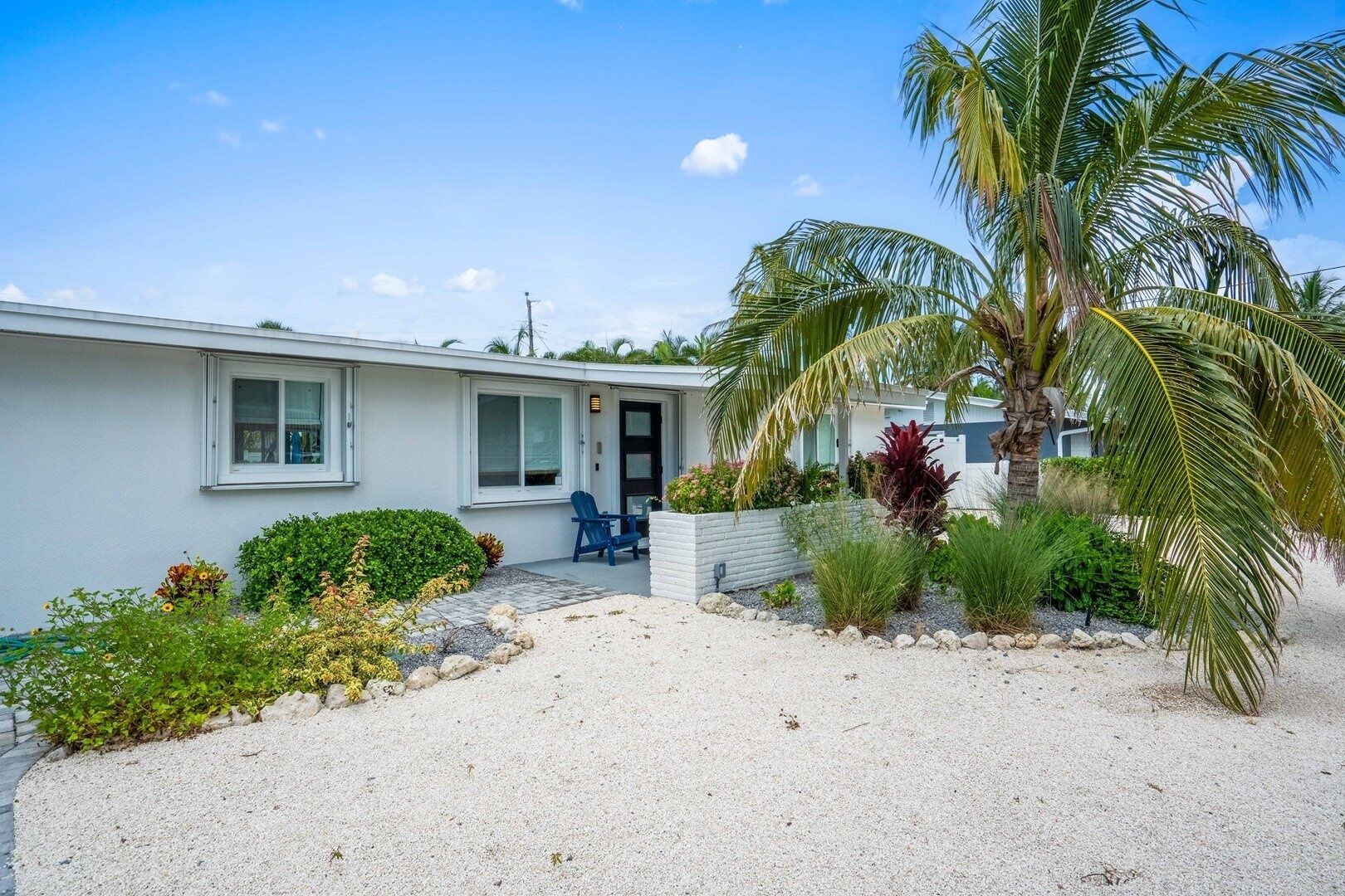 A single-story house with a front porch, a blue chair, and a palm tree.