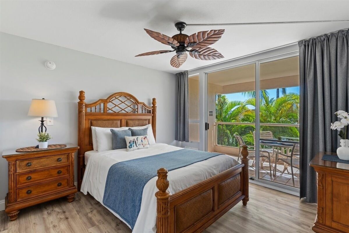 Een slaapkamer met een houten bed, een nachtkastje, een plafondventilator en uitzicht op een balkon met buitenmeubilair.