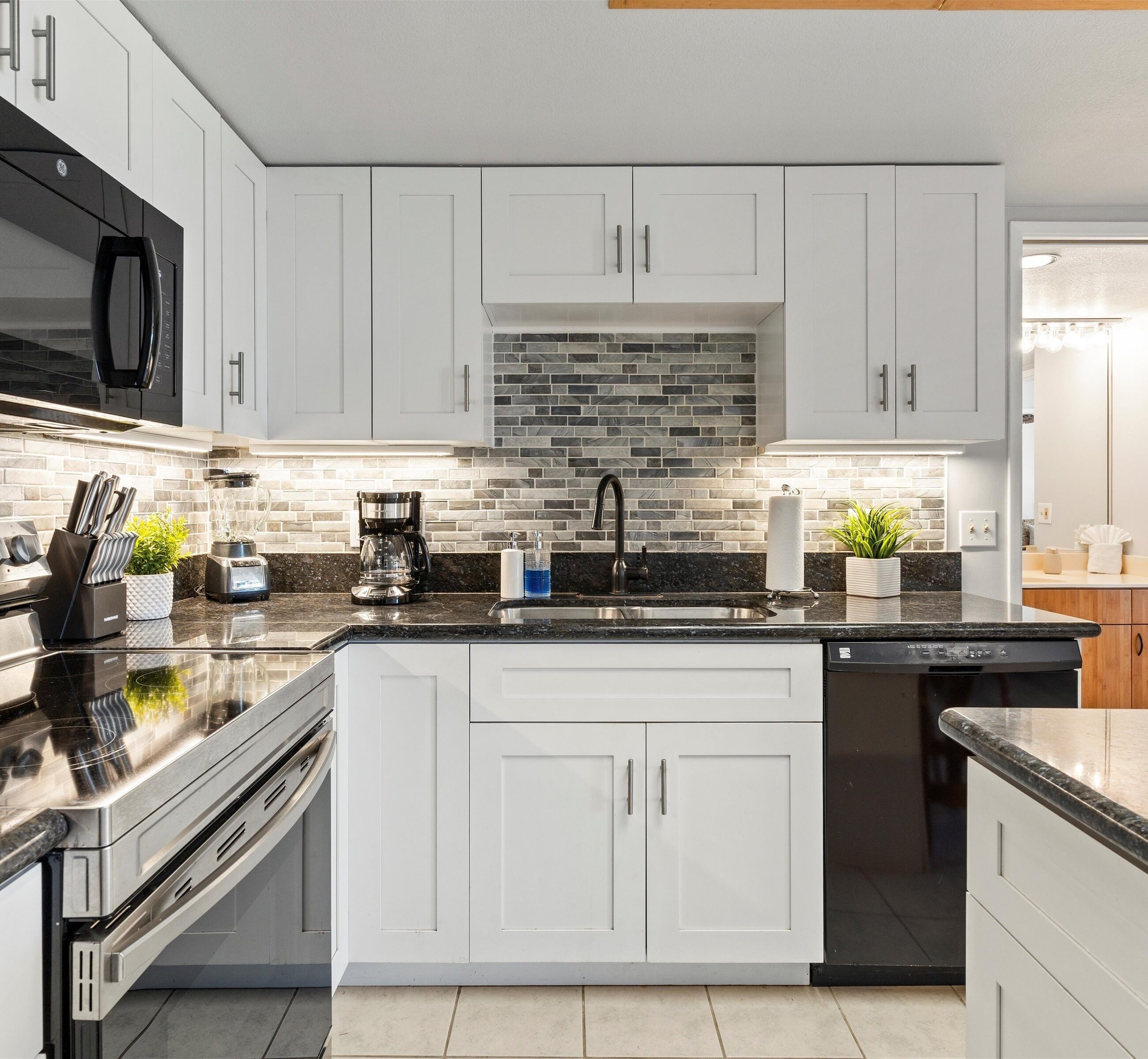 A modern kitchen with white cabinets, a black countertop, a brick backsplash, and a dishwasher.