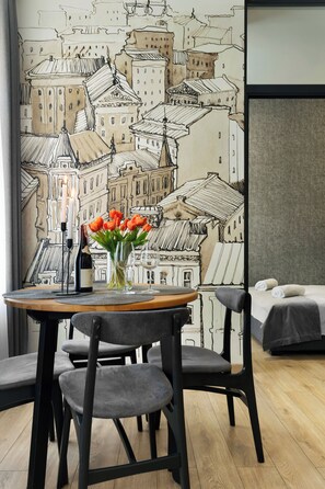 Habitación de hotel con zona de comedor que incluye una mesa dispuesta con una botella de vino, un jarrón de tulipanes y dos copas. La habitación tiene un mural grande con un paisaje urbano en la pared y una cama con una manta gris.
