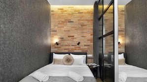Een moderne hotelkamer met twee bedden, grijze lakens, een bakstenen muur en een glazen scheidingswand.
