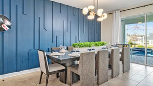 Een moderne eetruimte met een blauwe accentmuur, een grote tafel gedekt voor zes personen en uitzicht op een zwembad door grote ramen.