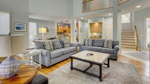 A spacious living area with a grey sofa set, a coffee table, and a dining table. There is a staircase leading to an upper level, and a kitchen area is visible in the background.