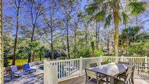 A deck with a table and chairs, surrounded by a white railing, overlooking a lake and dense trees.