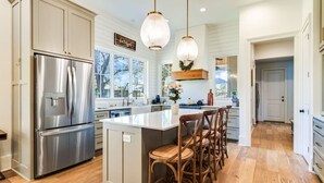 Una cocina moderna con refrigerador de acero inoxidable, isla de madera con taburetes altos e iluminación colgante, y vistas al exterior.