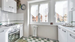 Une cuisine avec des meubles blancs, une cuisinière à gaz, un sol carrelé et une fenêtre donnant sur des immeubles.