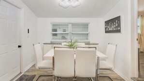 A dining area with a white table, four chairs, a potted plant, and a window with blinds.