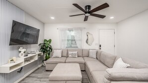 A modern living room with a sectional sofa, a flat-screen TV mounted on the wall, a ceiling fan, and a potted plant.