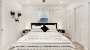 A bedroom with a bed, a ceiling fan, a window with curtains, bedside tables with lamps, and a wardrobe.