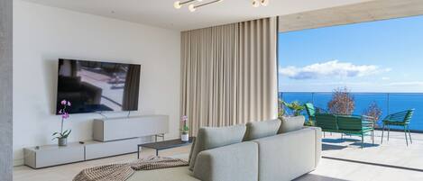 A modern living room with a large flat-screen TV, a beige sofa, and a balcony with green chairs overlooking a body of water.