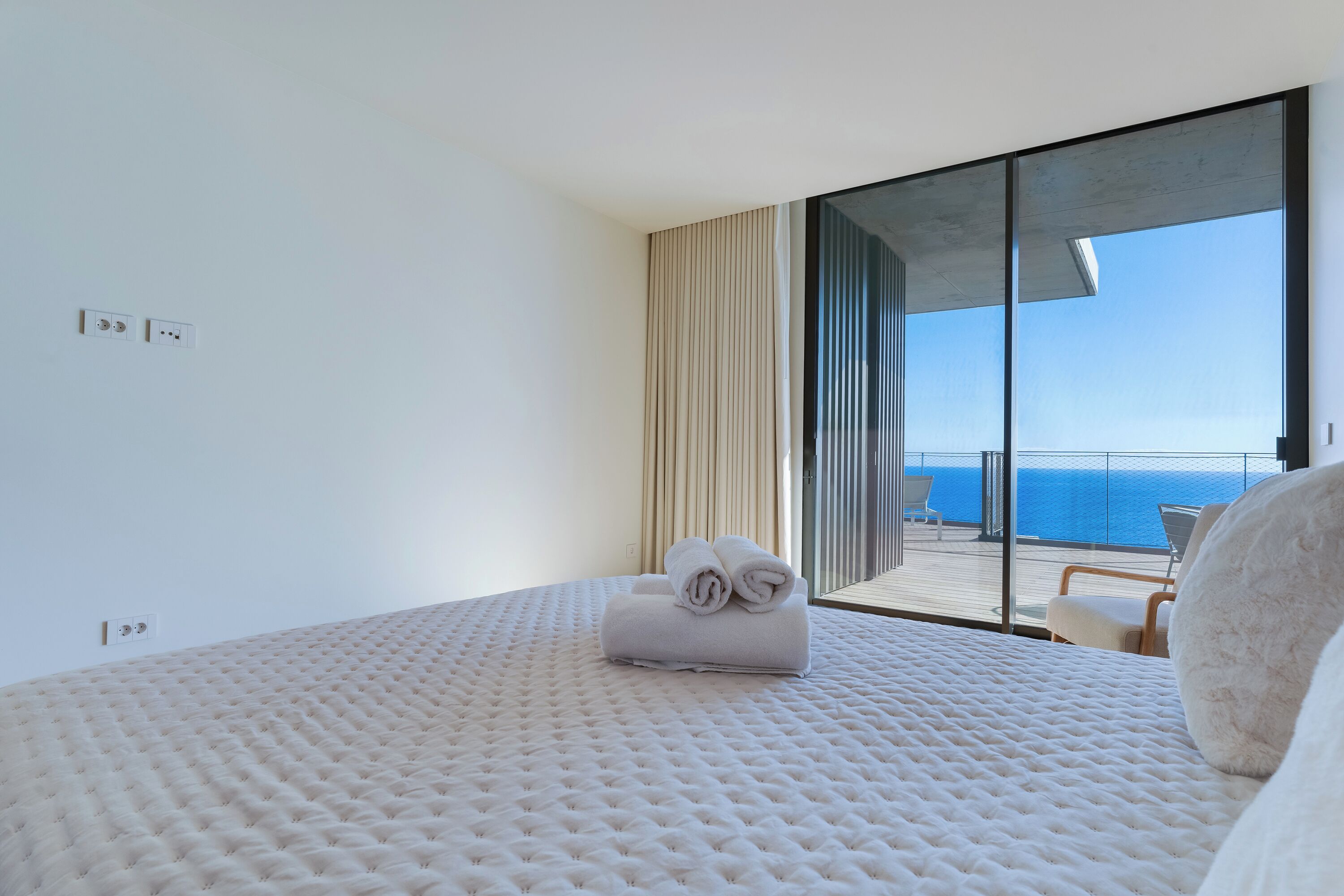 A bedroom with a large bed, a view of the ocean, and a balcony with a chair.