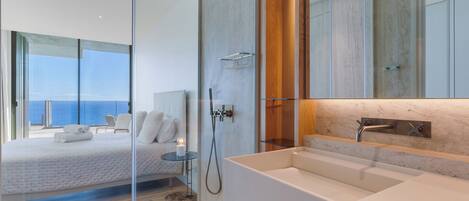 A modern bedroom with a large bed, a glass shower enclosure, and a view of the sea.