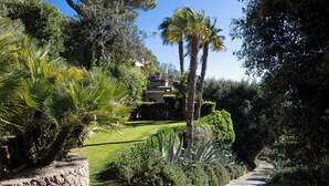 Un sendero de piedra serpentea por un exuberante jardín con palmeras y diversos arbustos. Se divisa un edificio con techo de tejas al fondo.