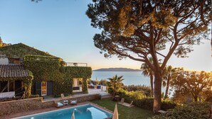 Zona de piscina con sillas de descanso y sombrillas, un muro de piedra y vista al mar.
