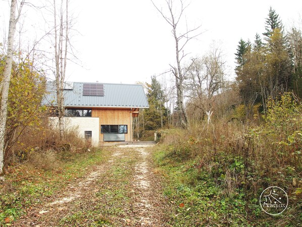 Una casa moderna con tetto in metallo e facciata in legno, immersa in una fitta foresta e un sentiero sterrato che conduce all'ingresso.