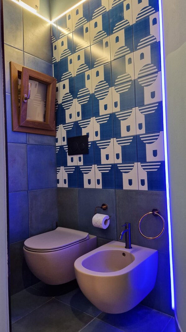 A bathroom with a toilet and bidet, tiled walls, a framed picture, and a toilet paper holder.