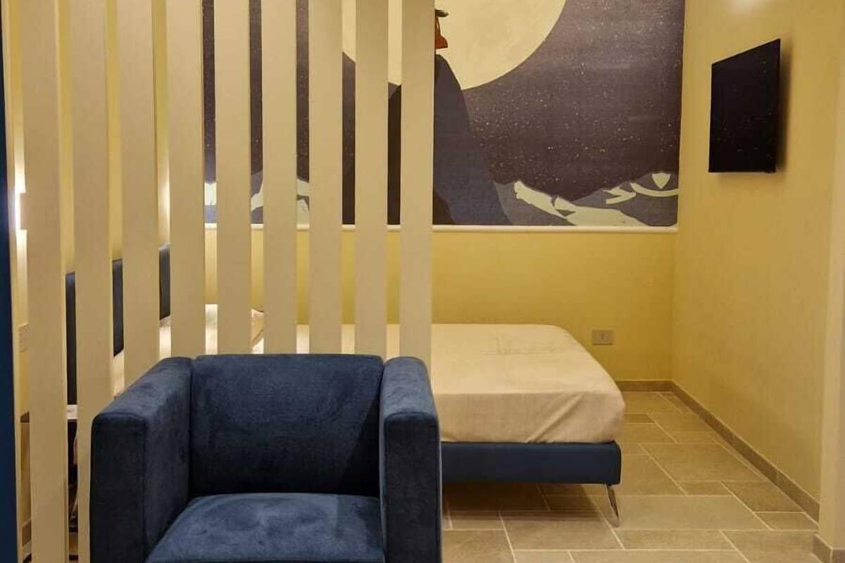 A room with a blue sofa, a bed, a large moon painting on the wall, and a TV mounted on the wall.