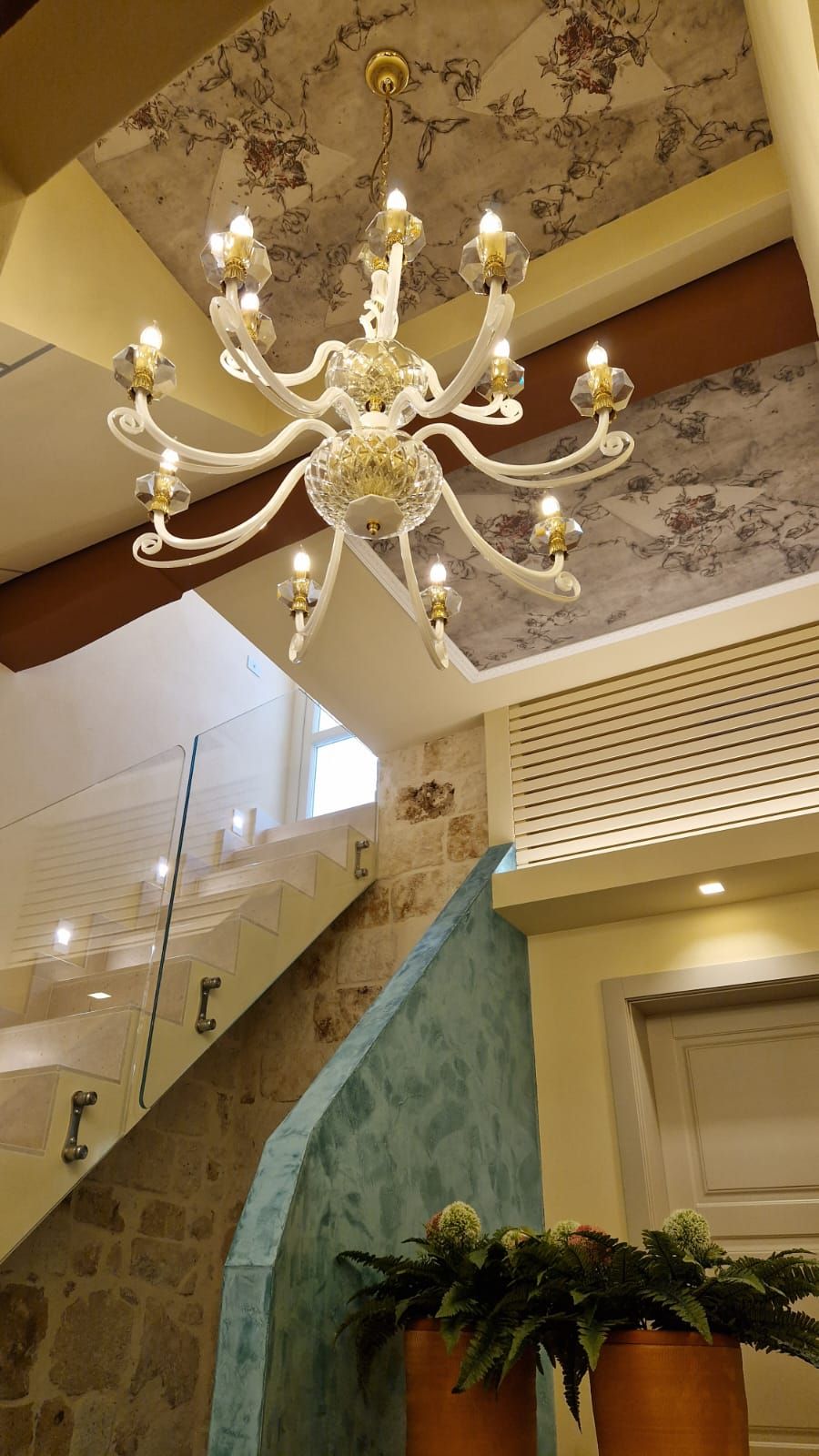 Un lampadario con molteplici bracci e luci, una scala con parete di fondo azzurra e piante in vaso alla base.