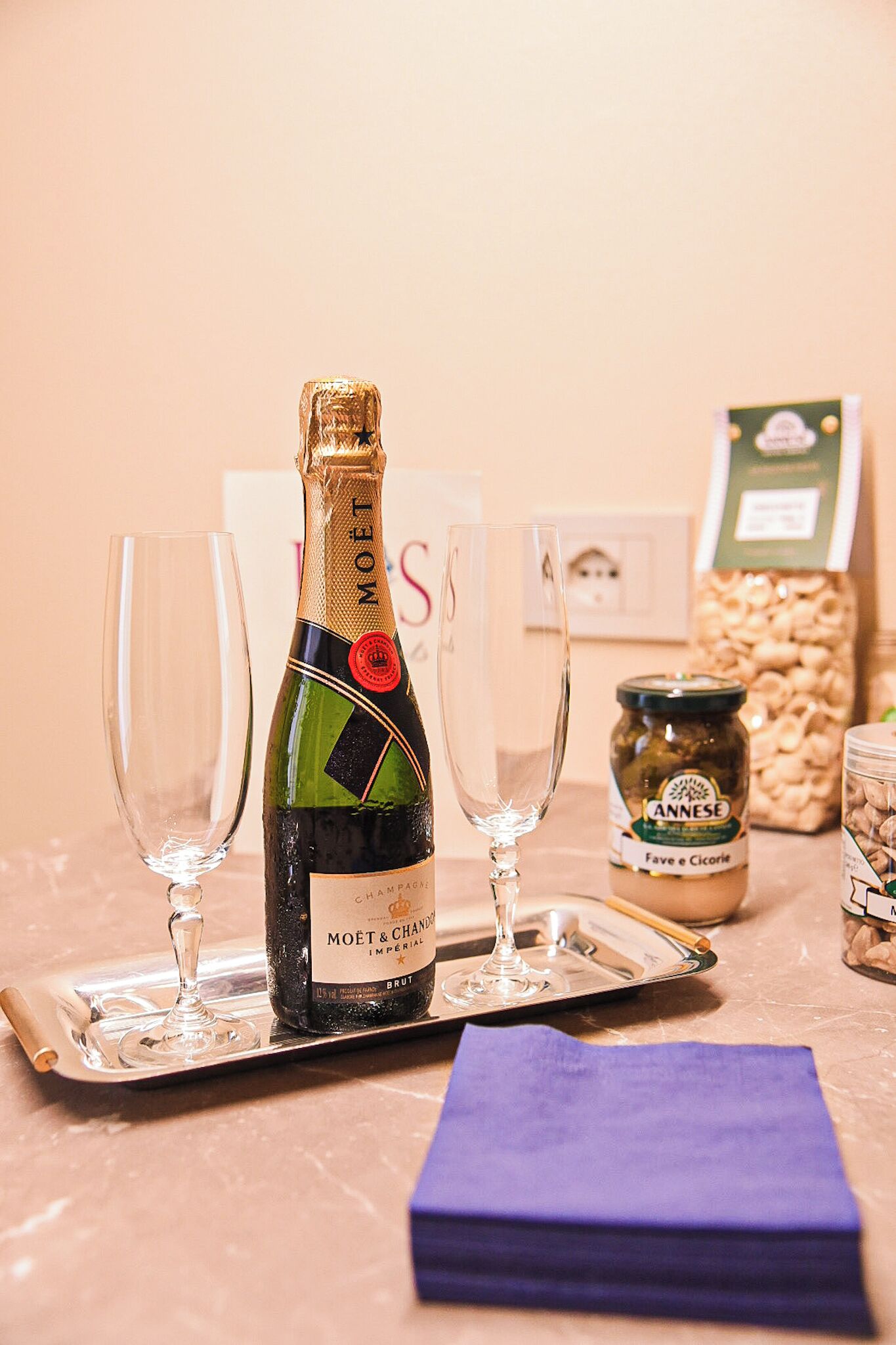 Eine Flasche Moët & Chandon Champagner auf einem Tablett mit zwei leeren Gläsern, ein Glas ANNESE Oliven und ein violettes Servietttuch auf einem Marmortisch.