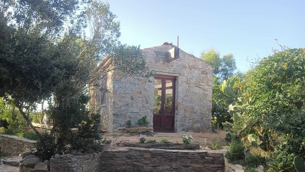 Un edificio de piedra con una puerta roja, rodeado de follaje y un muro de piedra.