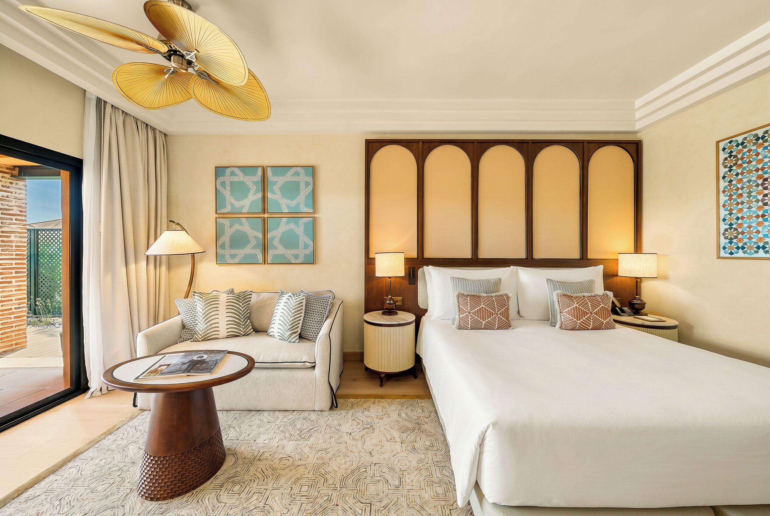 A hotel room with a bed, a sofa, a coffee table, a floor lamp, a wall-mounted fan, and artwork.