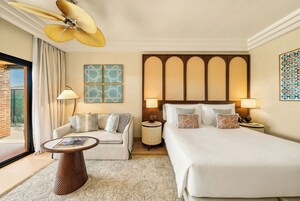 A hotel room with a bed, a sofa, a coffee table, a floor lamp, a wall-mounted fan, and artwork.
