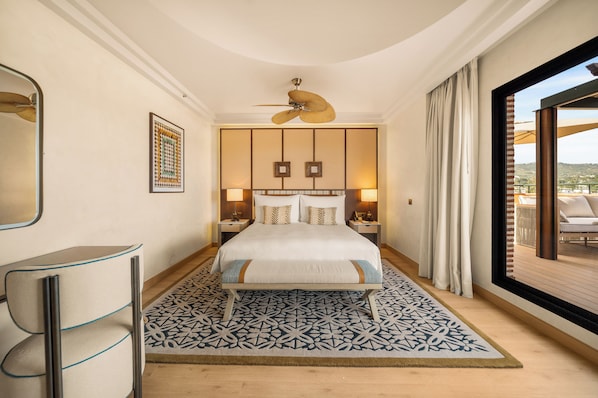 A hotel room with a bed, a ceiling fan, a large window with curtains, a patterned rug, and a nightstand with a lamp. - Gran Marbella Resort & Beach Club (Marbella)