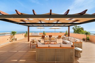 A wooden deck with outdoor seating, umbrellas, and a view of the sea.