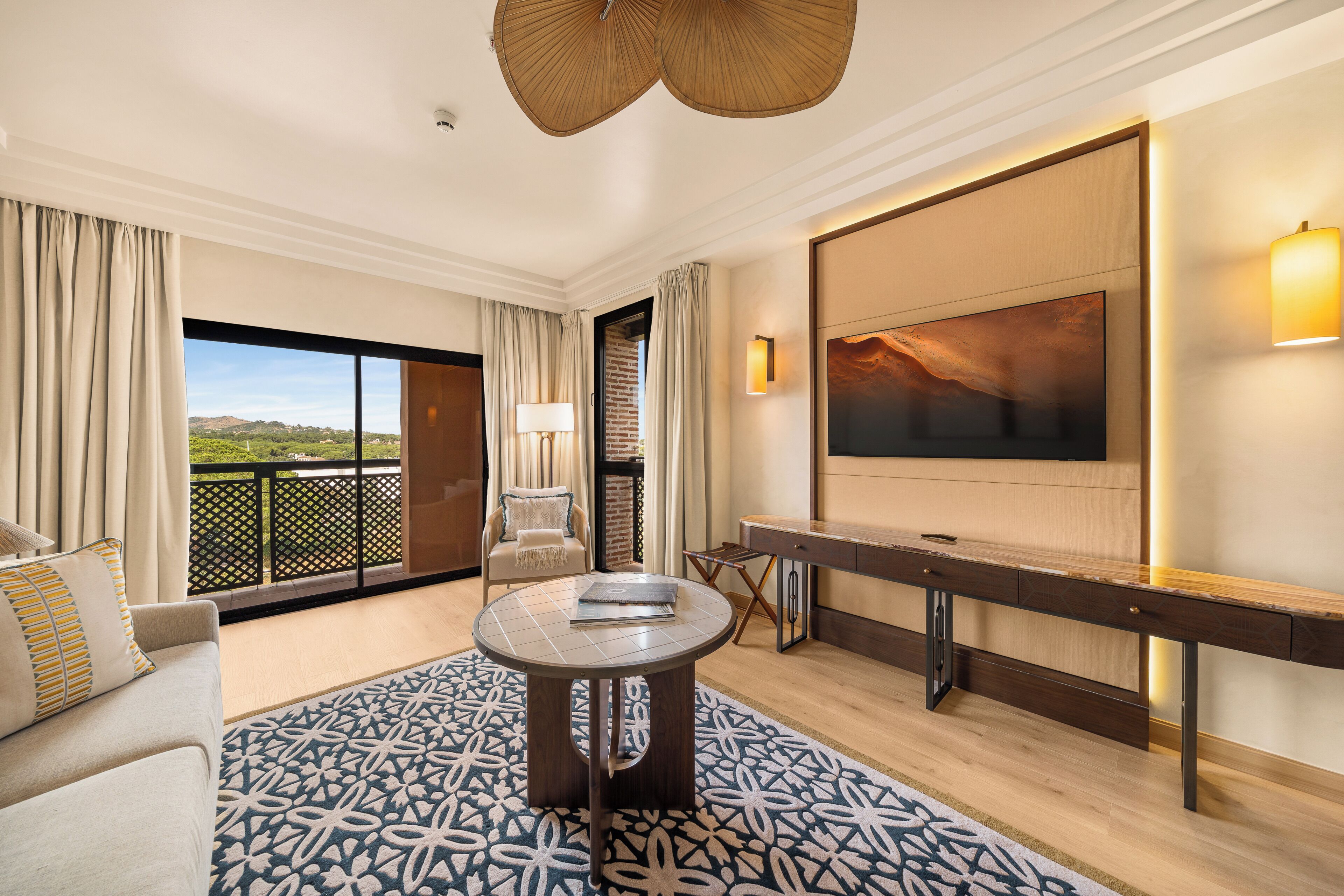 Een hotelkamer met balkon, een ronde tafel, een bank, een bureau en een schilderij aan de muur.