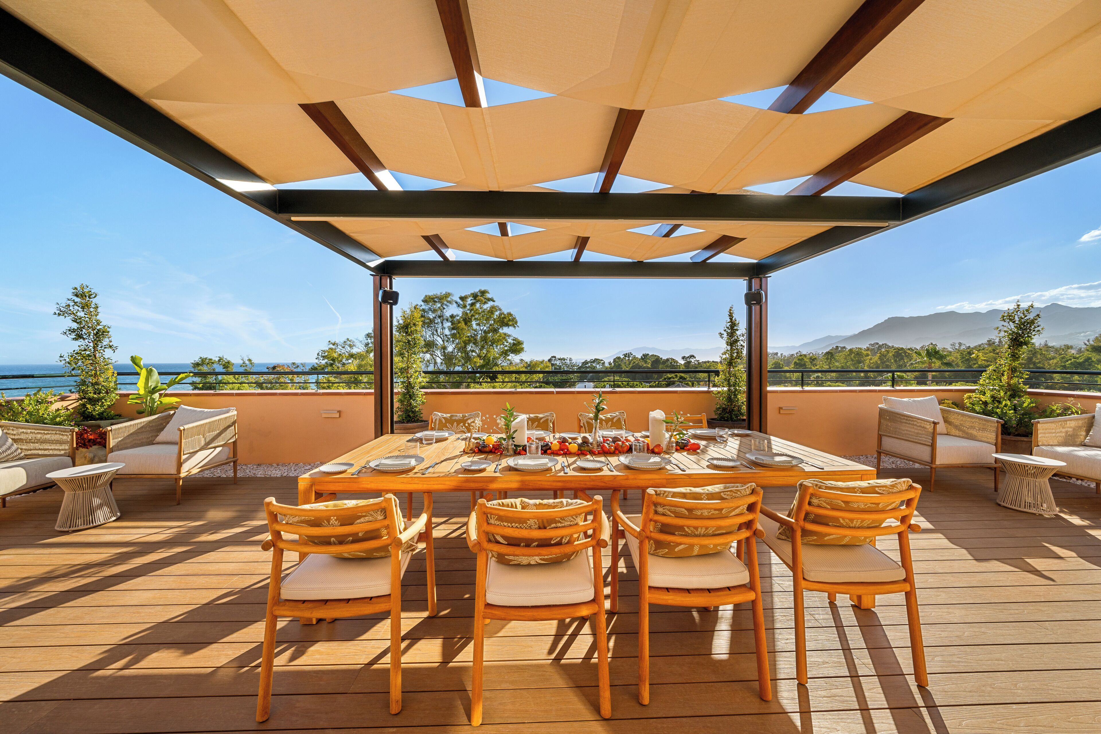 An outdoor dining area with a table set for six, surrounded by wicker chairs, under a shaded pergola.