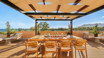 Un área de comedor al aire libre con una mesa dispuesta para seis personas, rodeada de sillas de mimbre, bajo una pérgola sombreada.