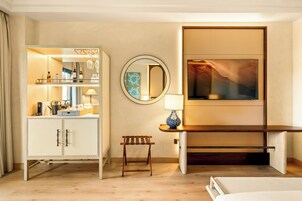A hotel room with a minibar, a round mirror, a bench, and a painting on the wall.