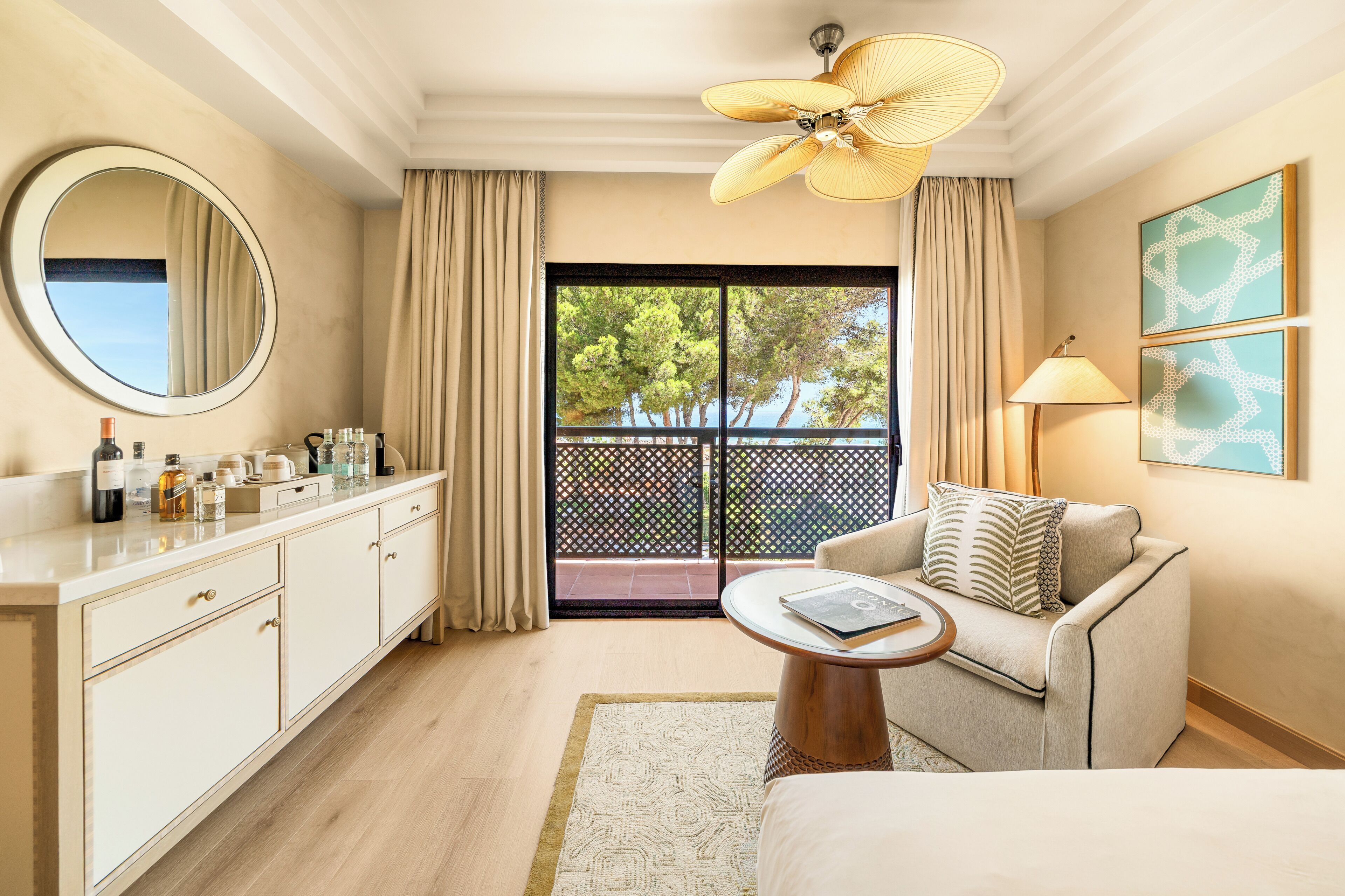 A hotel room with a balcony, a round mirror, a sofa, a coffee table, and a ceiling fan.