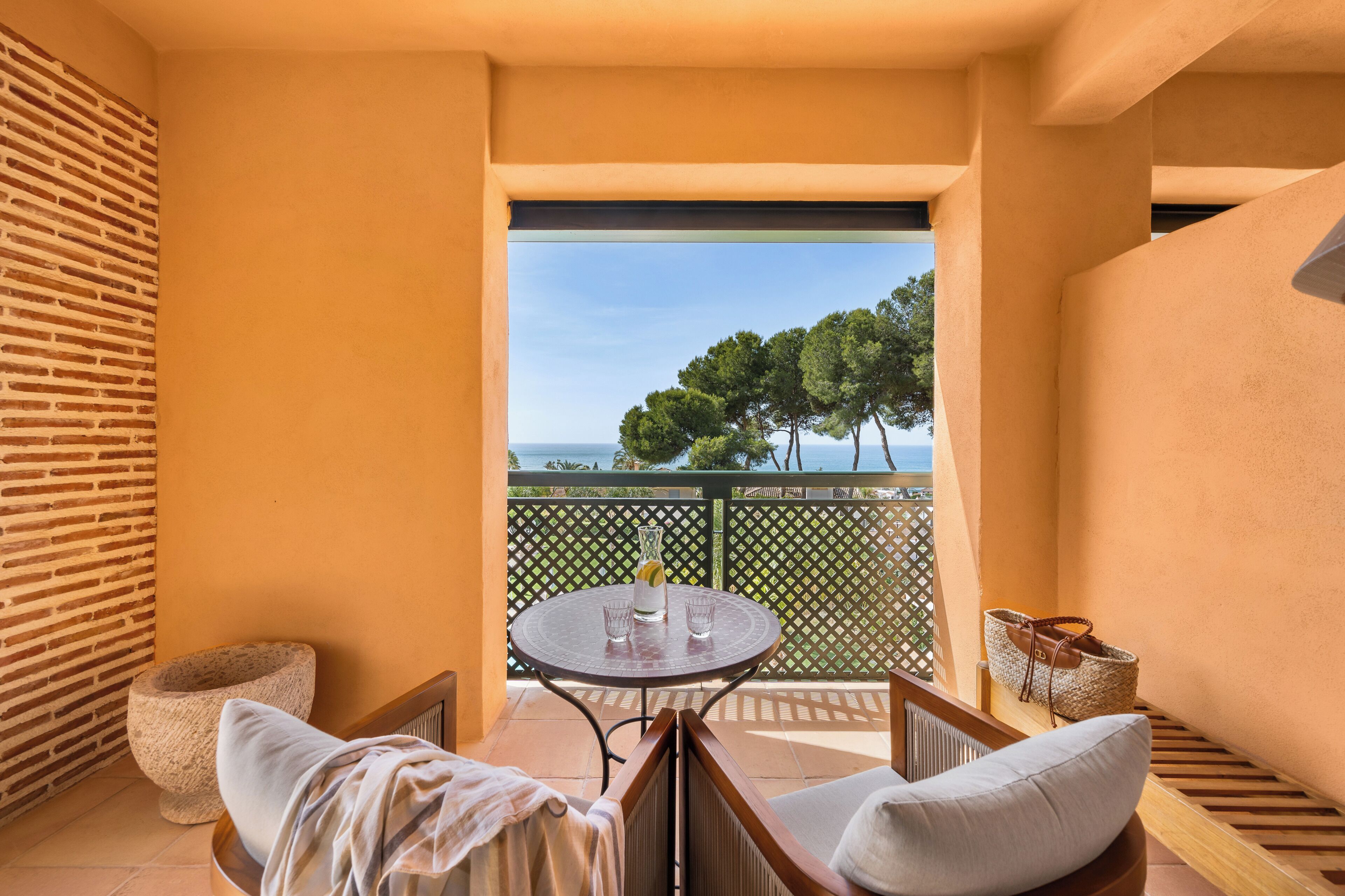 A balcony with a table, chairs, and a view of trees and the sea.