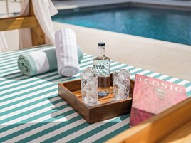 Een zwembadterras met een gestreepte handdoek, een fles Gran Reserva, twee glazen met ijs en een boek getiteld 'ICONIC'.