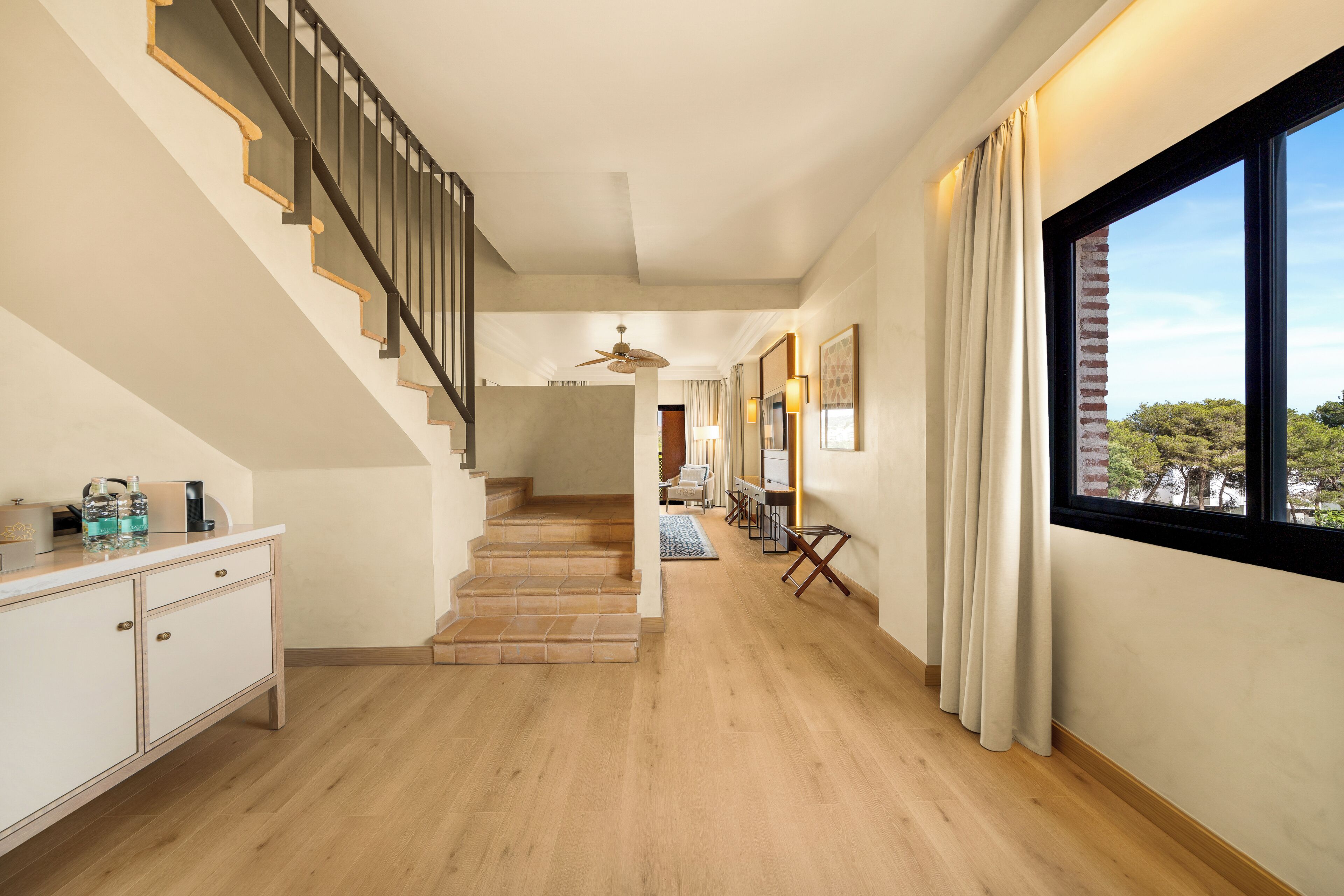 Passadís d'hotel amb escala, sòl de fusta, escriptori amb cadira i una finestra gran amb cortines.
