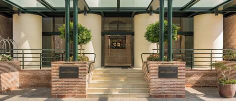 Entrada de hotel con toldo de vidrio, escalones de ladrillo y plantas en macetas.