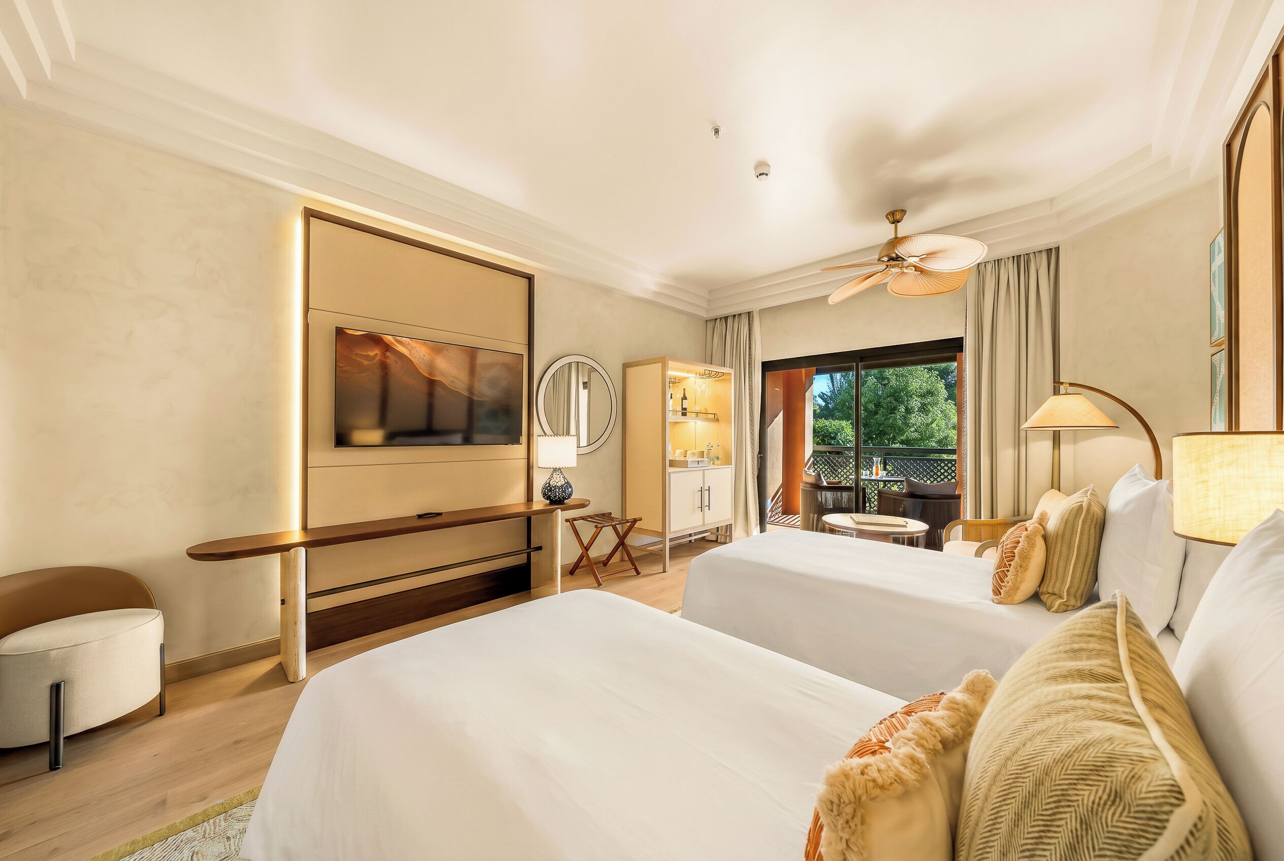 A hotel room with two beds, a TV, a desk, a chair, a ceiling fan, and a view of greenery outside.