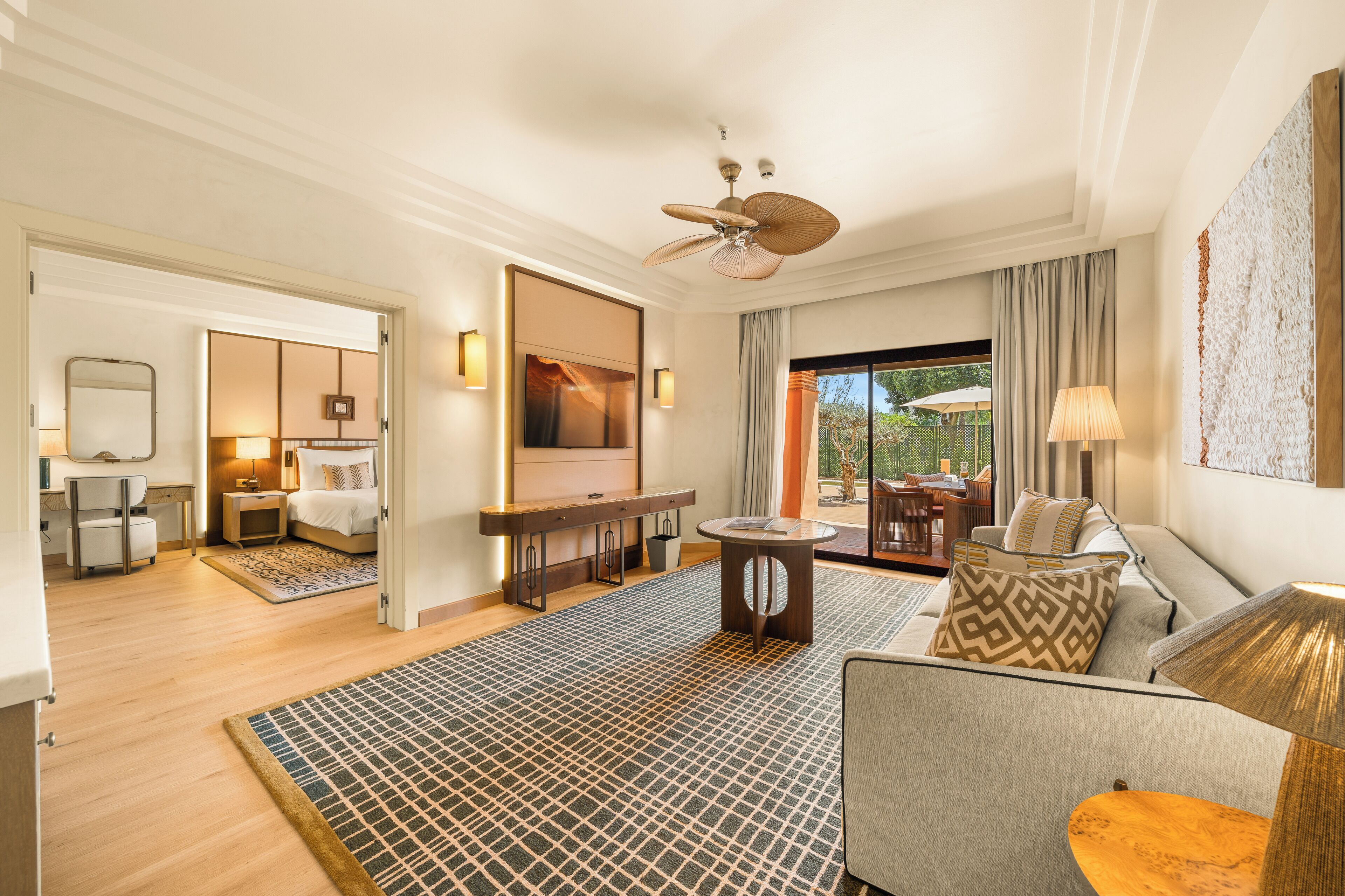 Habitación de hotel con cama, zona de estar con sofá, mesa de comedor, televisor montado en la pared y vistas al exterior.