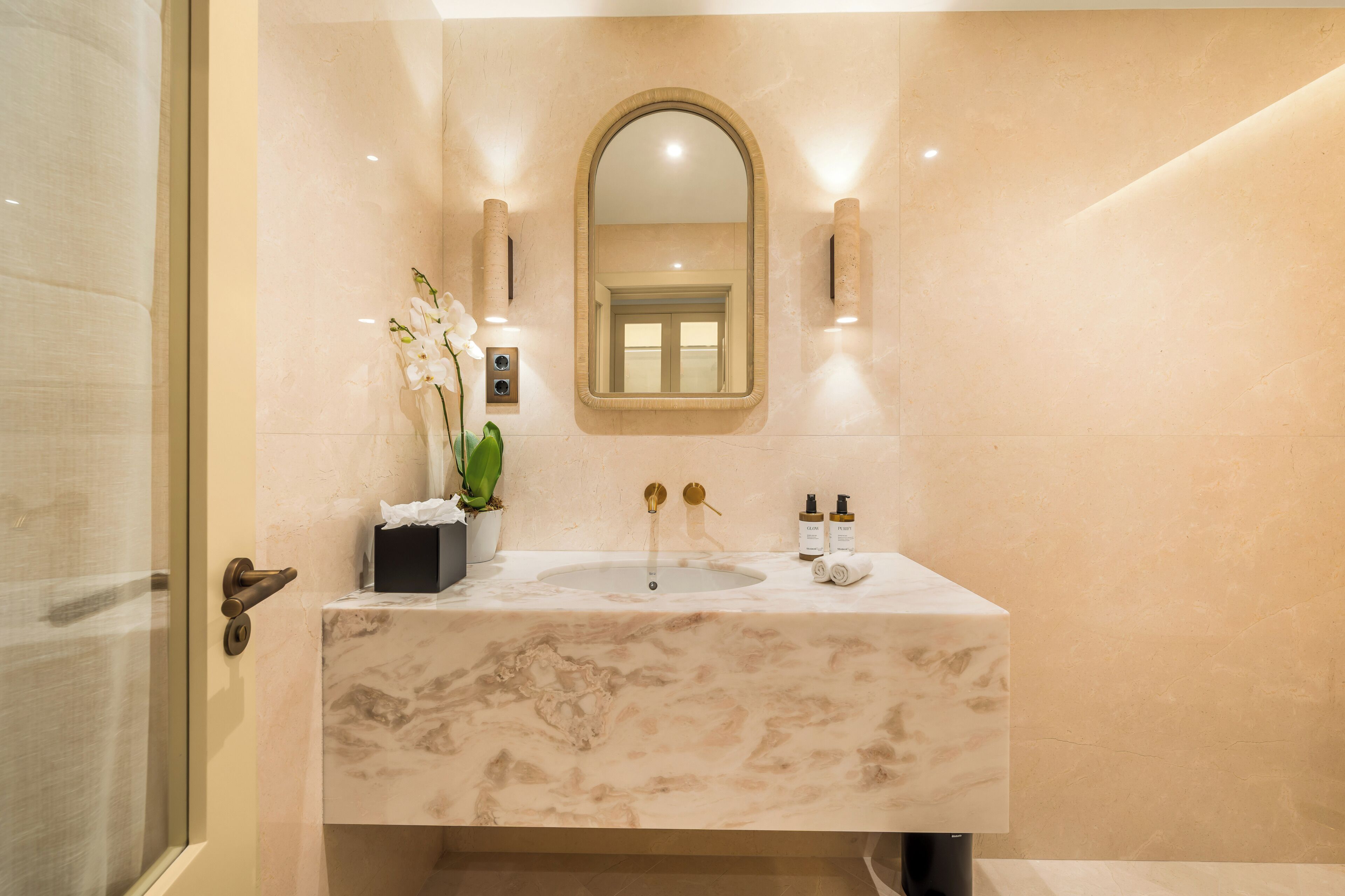 A bathroom with a marble sink, a mirror, a potted orchid, and two bottles of toiletries.