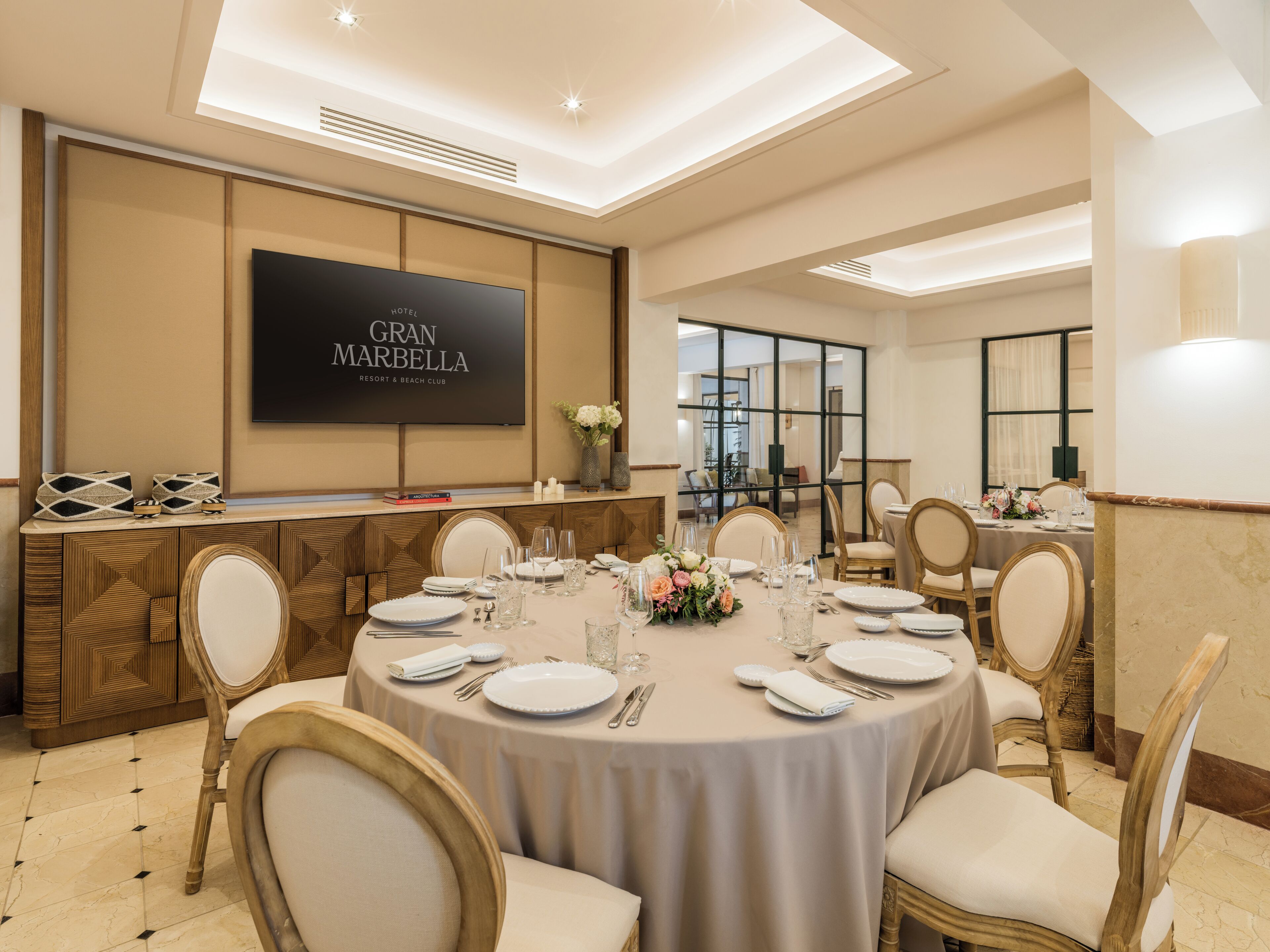 Un comedor de hotel con mesas redondas dispuestas para el servicio, una pantalla grande mostrando 'GRAN MARBELLA RESORT & BEACH CLUB', y vista al salón comedor.