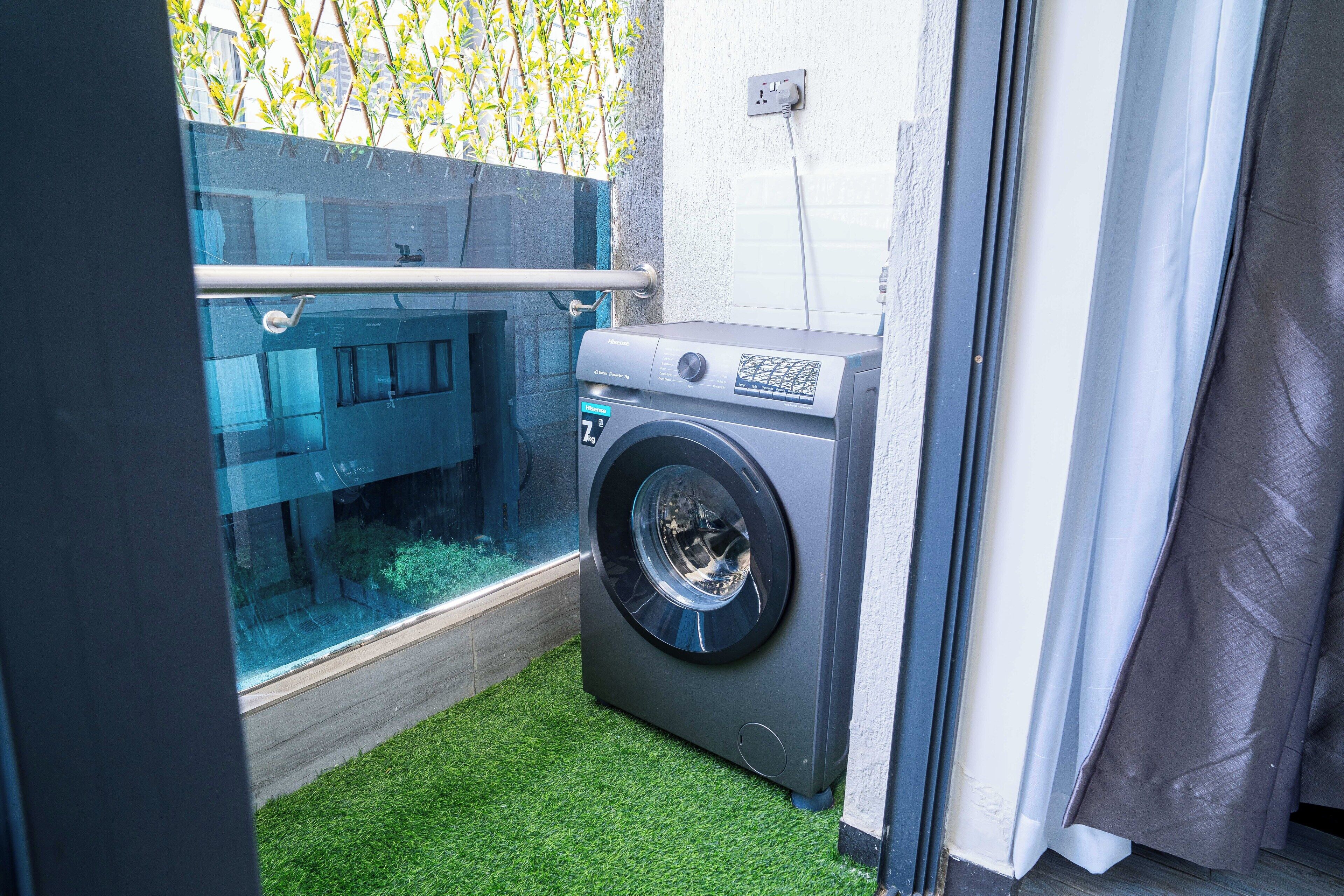 Un espace réduit dédié à la buanderie, comprenant un lave-linge, une petite fenêtre et un sol recouvert d’un gazon synthétique vert.