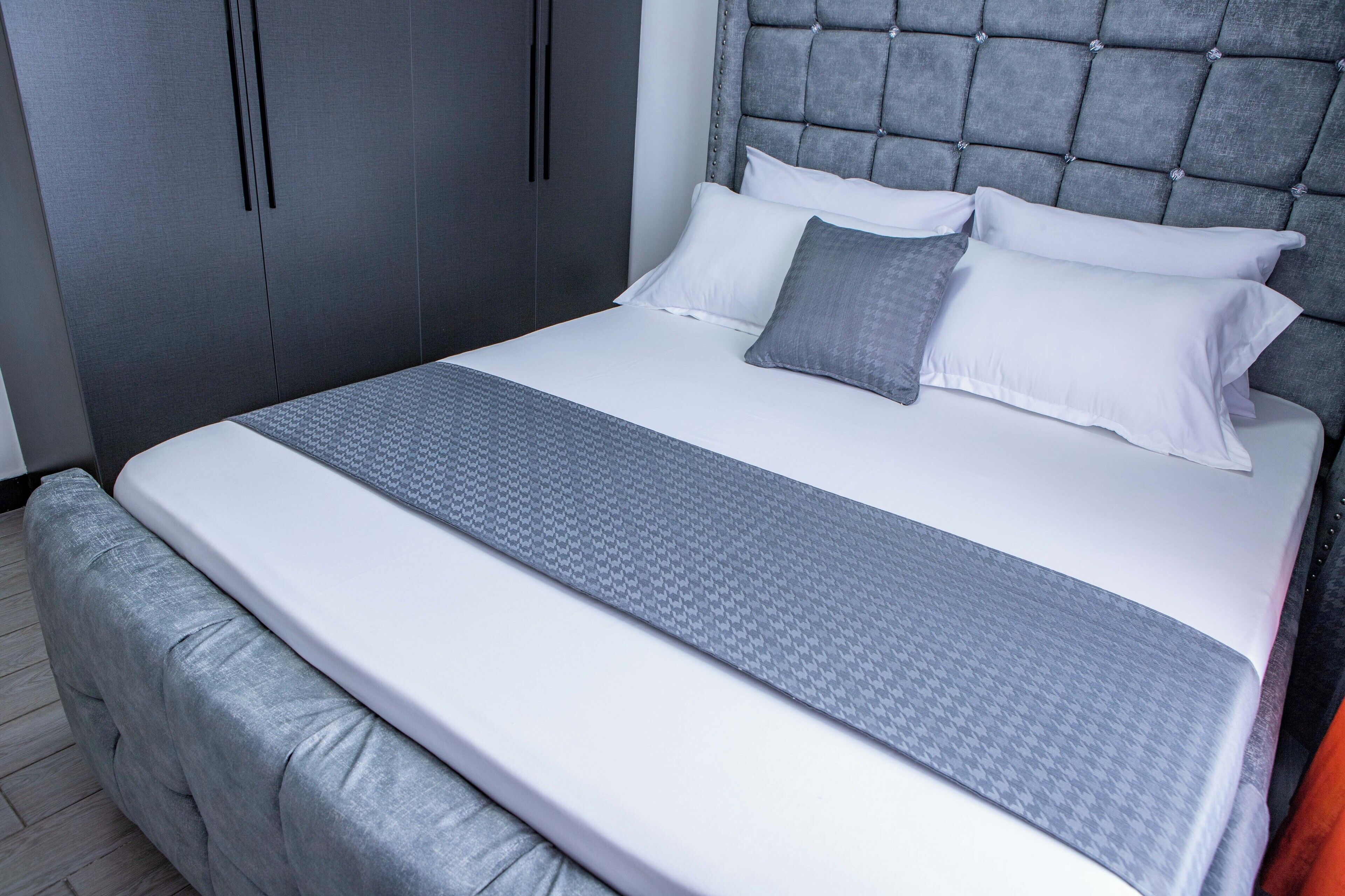 Una cama bien hecha con un edredón gris estampado y almohadas, un cabecero tapizado en gris y un armario gris con asas verticales.