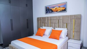 A neatly made bed with white and orange bedding, a grey wardrobe, and a white bedside table with a heater. - Maskan Airbnbs (Nairobi)