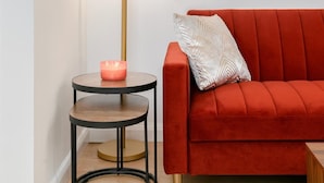A red sofa with a white patterned pillow, a round side table with a candle, and a floor lamp. - Deluxe 1 Bed Apartment, London (London)