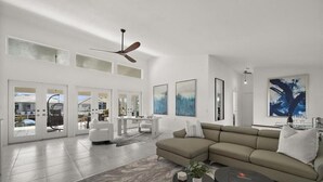 Un salon spacieux avec un grand canapé d’angle, une table basse et un ventilateur de plafond. De grandes fenêtres et des portes donnent sur un espace extérieur.